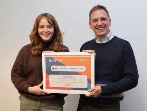 Two people smiling and holding a framed ECPO Award 2024 certificate against a plain background.