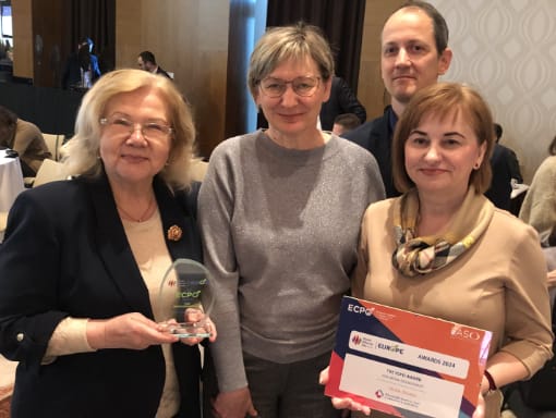 Four people are in an indoor setting, two women hold an award and a certificate, respectively. They are dressed in business-casual attire.