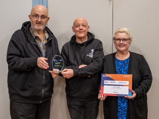 Three people stand together indoors. The person in the middle holds a glass award, and the person on the right holds a certificate. All are smiling.